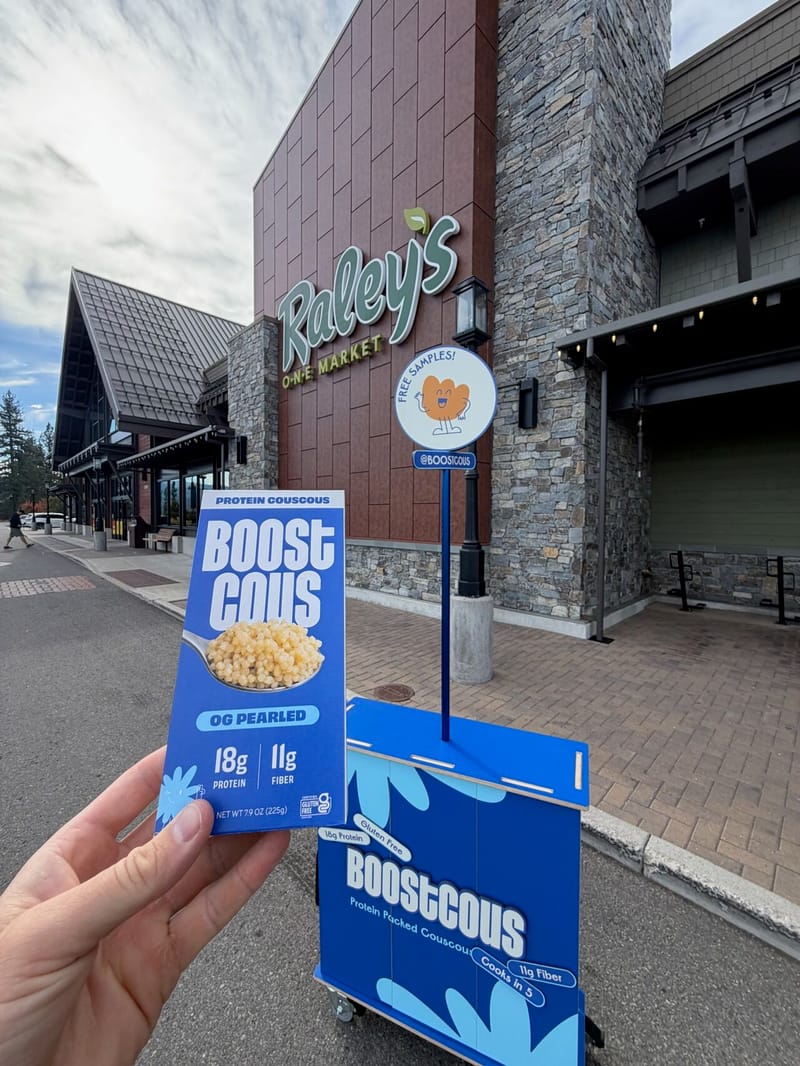 A hand holding a Boostcous box in front of a Raley's O-N-E Market store with a blue Boostcous sampling cart and Free Samples sign outside